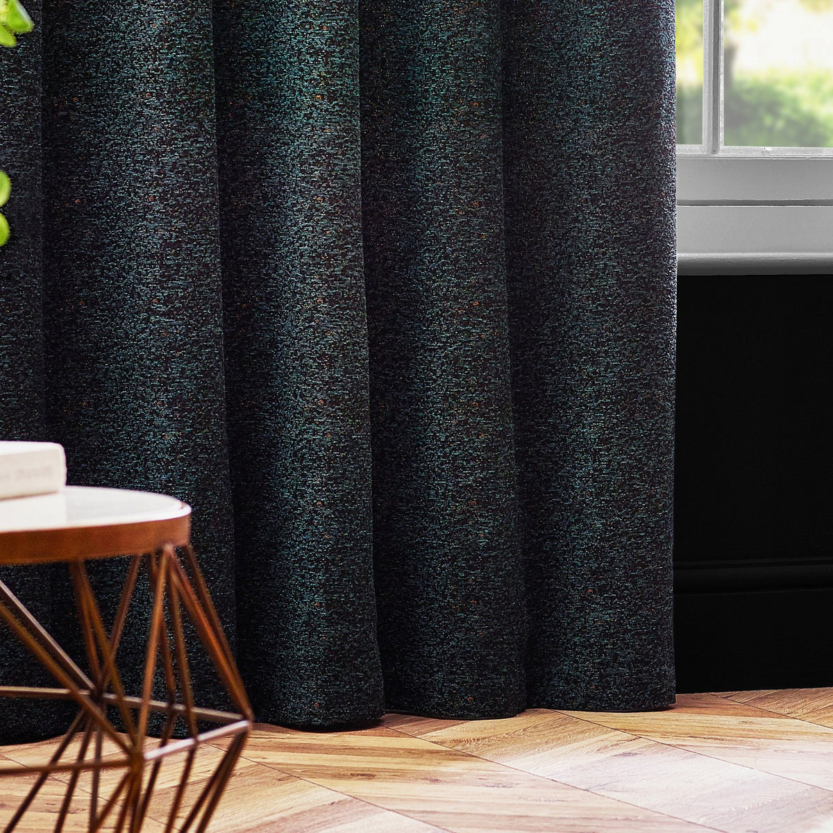 Galaxy Chenille Fully Lined Eyelet Curtains in Emerald, shown in soft folds by a window in a living room with wooden flooring and a round side table.