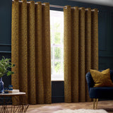 Galaxy Chenille Fully Lined Eyelet Curtains in gold, mottled textured fabric hung from a rail in a dark-walled living room with a matching cushion on a navy sofa.