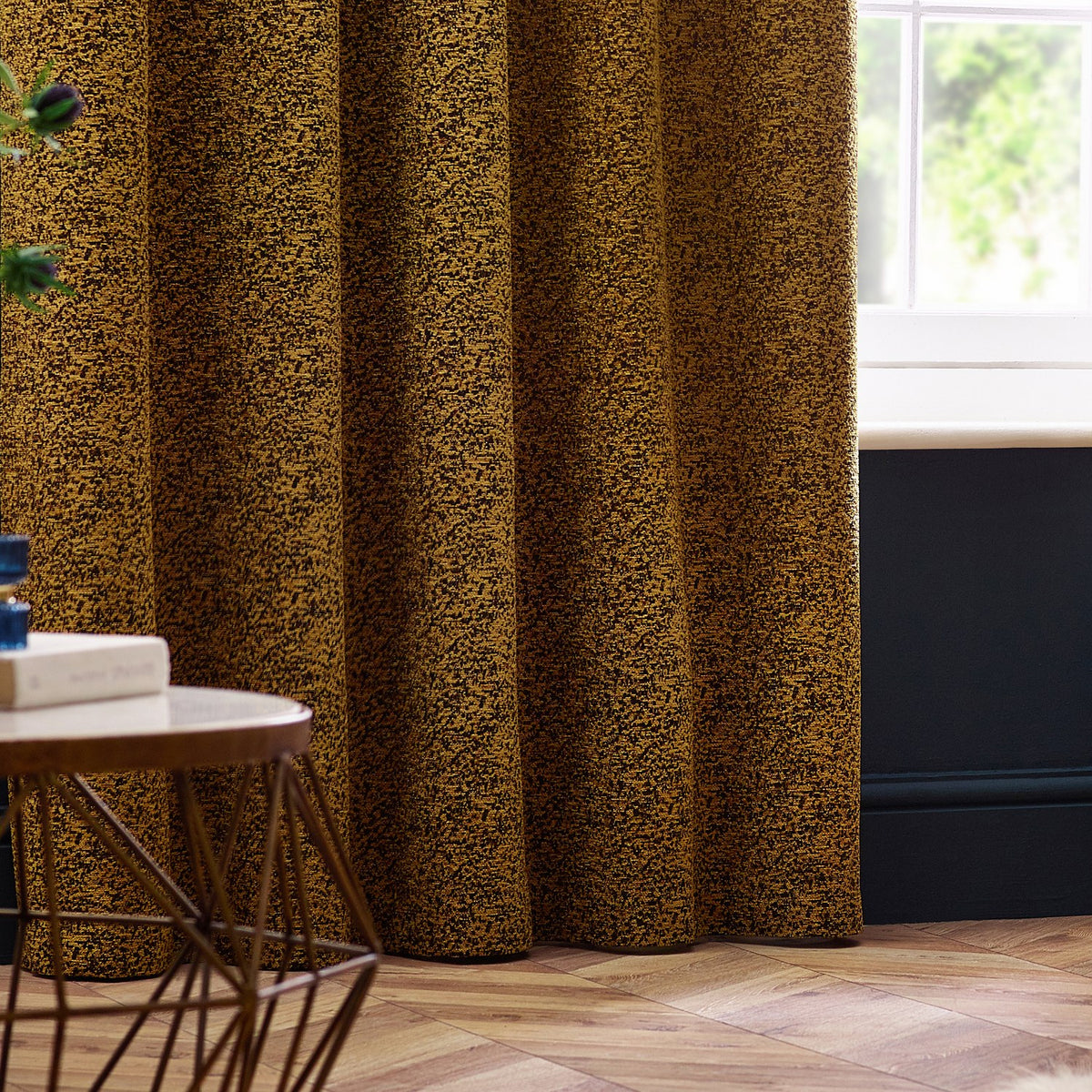 Galaxy fully lined eyelet curtains in gold chenille with a mottled woven texture, shown hanging in soft folds in a living room with wooden flooring.