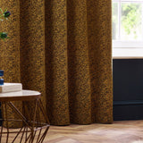Galaxy fully lined eyelet curtains in gold chenille with a mottled woven texture, shown hanging in soft folds in a living room with wooden flooring.