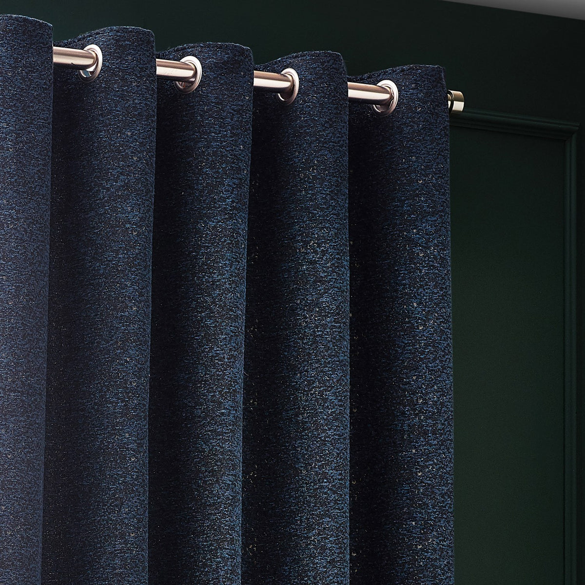 Galaxy Chenille Fully Lined Eyelet Curtains in navy, shown close-up on a brushed metal pole with eyelet rings, revealing a mottled chenille texture flecked with gold against a dark green wall.