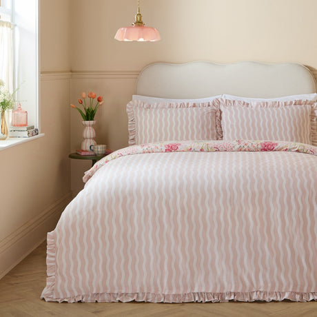 Gardenia Reversible Botanical Duvet Cover Set in Pink Pebble on a bed, showing the wavy pink and white stripe side with frilled edges, styled in a pale pink bedroom.