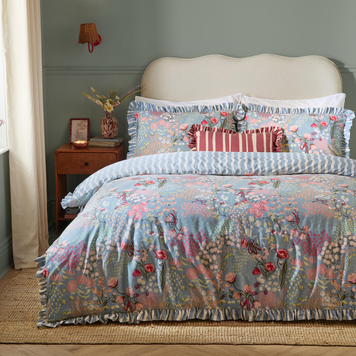 Gardenia Reversible Botanical Duvet Cover Set in Seaspray with ruffled edges and pink and coral floral print on blue-grey, styled with a red striped cushion and wooden nightstand.