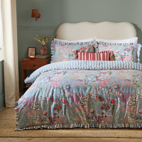 Gardenia Reversible Botanical Duvet Cover Set in Seaspray with ruffled edges and pink and coral floral print on blue-grey, styled with a red striped cushion and wooden nightstand.