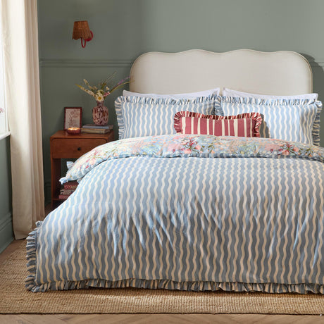 Gardenia Reversible Botanical Duvet Cover Set in Seaspray on a bed, wavy blue-cream striped side up with ruffled edges, floral reverse peeking through, striped bolster cushion, green-walled bedroom.