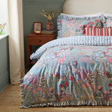 Gardenia Reversible Botanical Duvet Cover Set in Seaspray on a bed with ruffled edges, blue-grey floral print facing up, styled in a bedroom with a wooden nightstand.