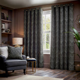 Gatsby Jacquard Lined Eyelet Curtains in black with champagne feathered arc pattern, hung by a window in a living room with matching cushion and armchair.