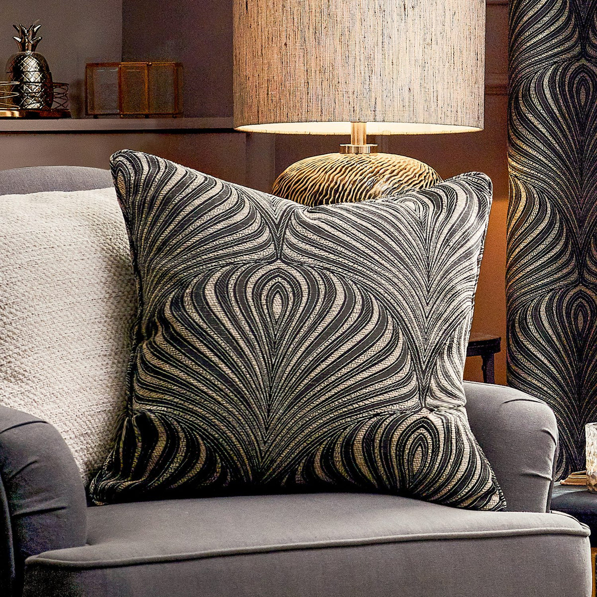 Gatsby Eyelet Polyester Cushion in black with woven Art Nouveau feather-like pattern in natural tone, shown on a grey armchair beside a table lamp.