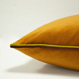 Close-up of the Gemini Reversible Velvet Cushion in pumpkin, showing amber-orange velvet with contrasting plum and lime double piping along the edge, on a plain light background.