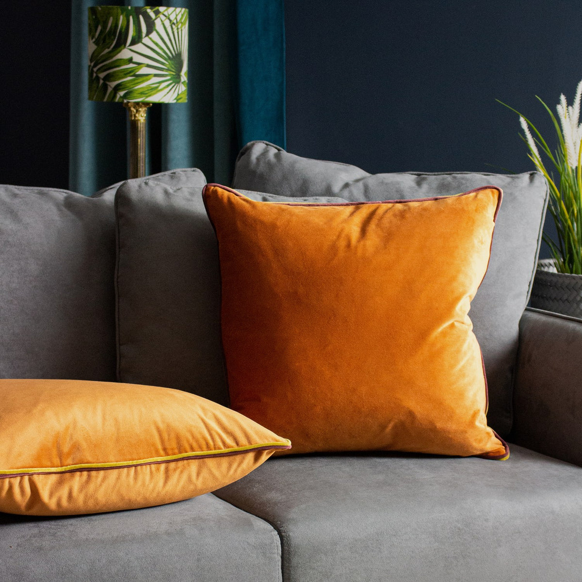 Gemini Reversible Velvet Cushion in pumpkin with contrast piping, shown on a grey sofa alongside a matching square velvet cushion in a living room setting.
