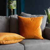 Gemini Reversible Velvet Cushion in pumpkin with contrast piping, shown on a grey sofa alongside a matching square velvet cushion in a living room setting.