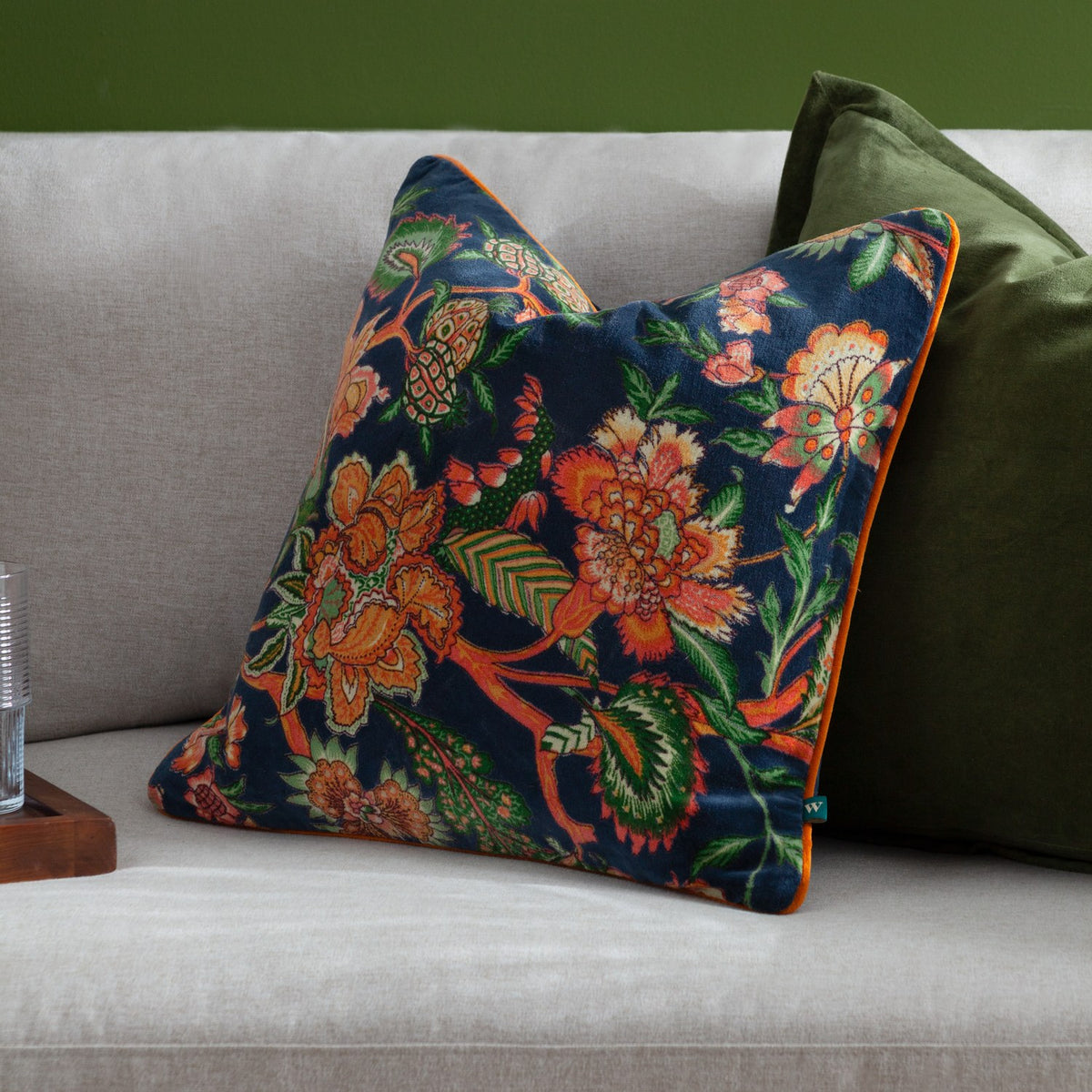 Georgia Velvet Cotton Cushion in royal blue with orange and green botanical print, orange piped edge, shown on a grey sofa beside a green cushion.