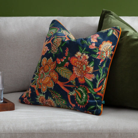 Georgia Velvet Cotton Cushion in royal blue with orange and green botanical print, orange piped edge, shown on a grey sofa beside a green cushion.