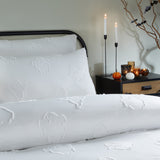 White tufted polyester duvet cover set with raised ghost motifs, shown on a bed beside a black nightstand with candles and pumpkins in a bedroom setting.