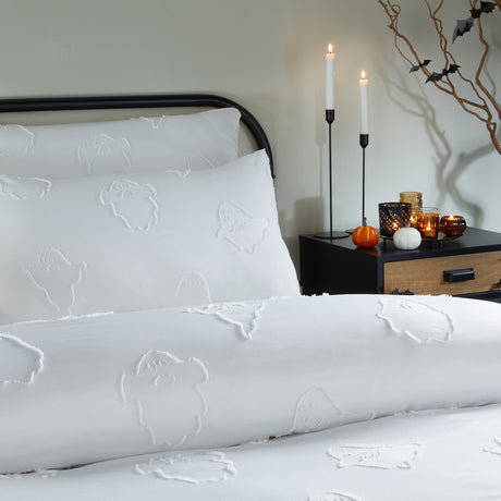 White tufted polyester duvet cover set with raised ghost motifs, shown on a bed beside a black nightstand with candles and pumpkins in a bedroom setting.