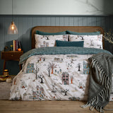 Ghost Town Reversible Woodland Duvet Cover Set in Dusk, cream-ground haunted house print with trees and pumpkins, styled on a bed with teal pillows in a bedroom setting.