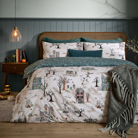 Ghost Town Reversible Woodland Duvet Cover Set in Dusk, cream-ground haunted house print with trees and pumpkins, styled on a bed with teal pillows in a bedroom setting.
