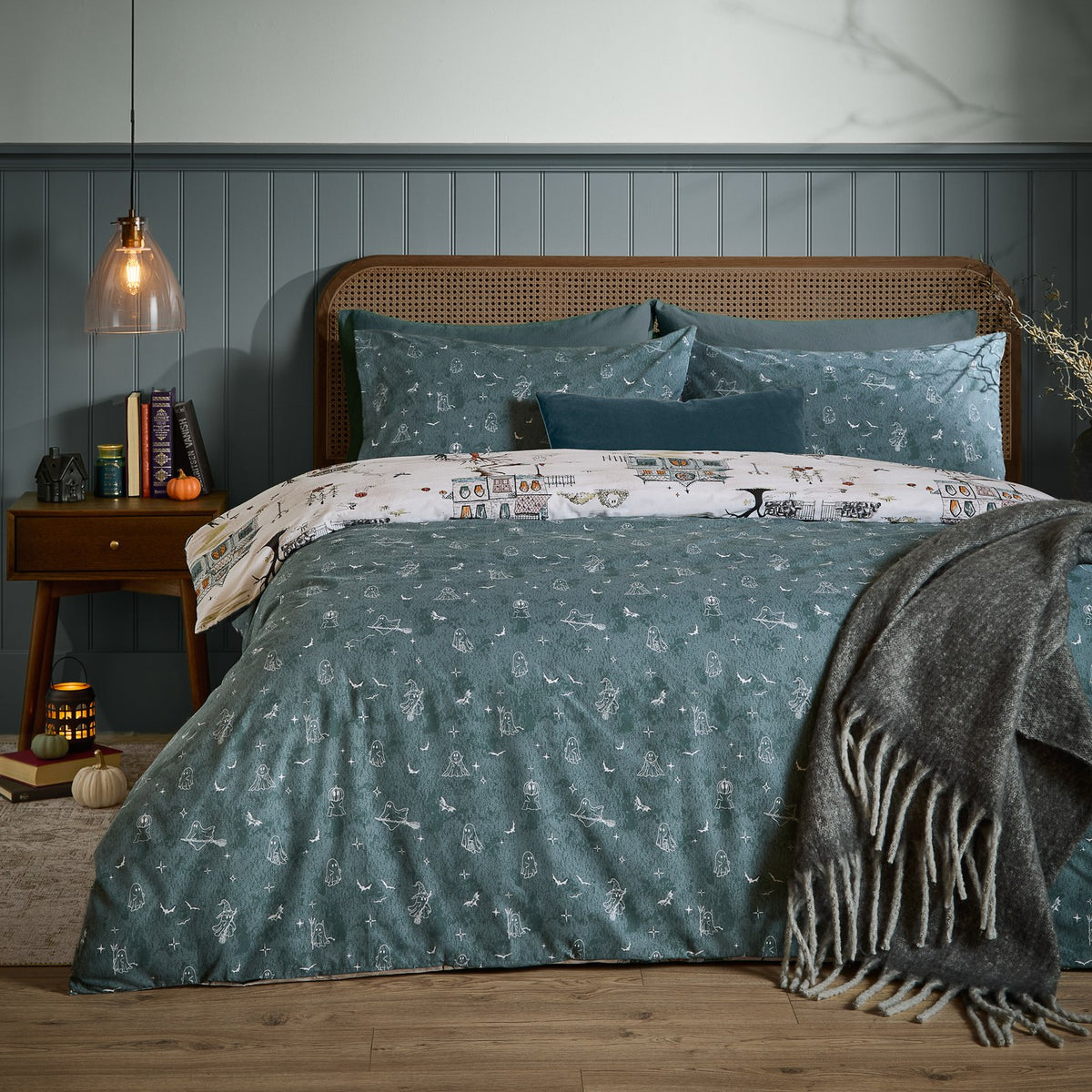 Ghost Town Reversible Woodland Duvet Cover Set in Dusk blue on a bed with cane headboard, showing cream ghost-motif reverse peeking at the pillows, styled in a moody bedroom.