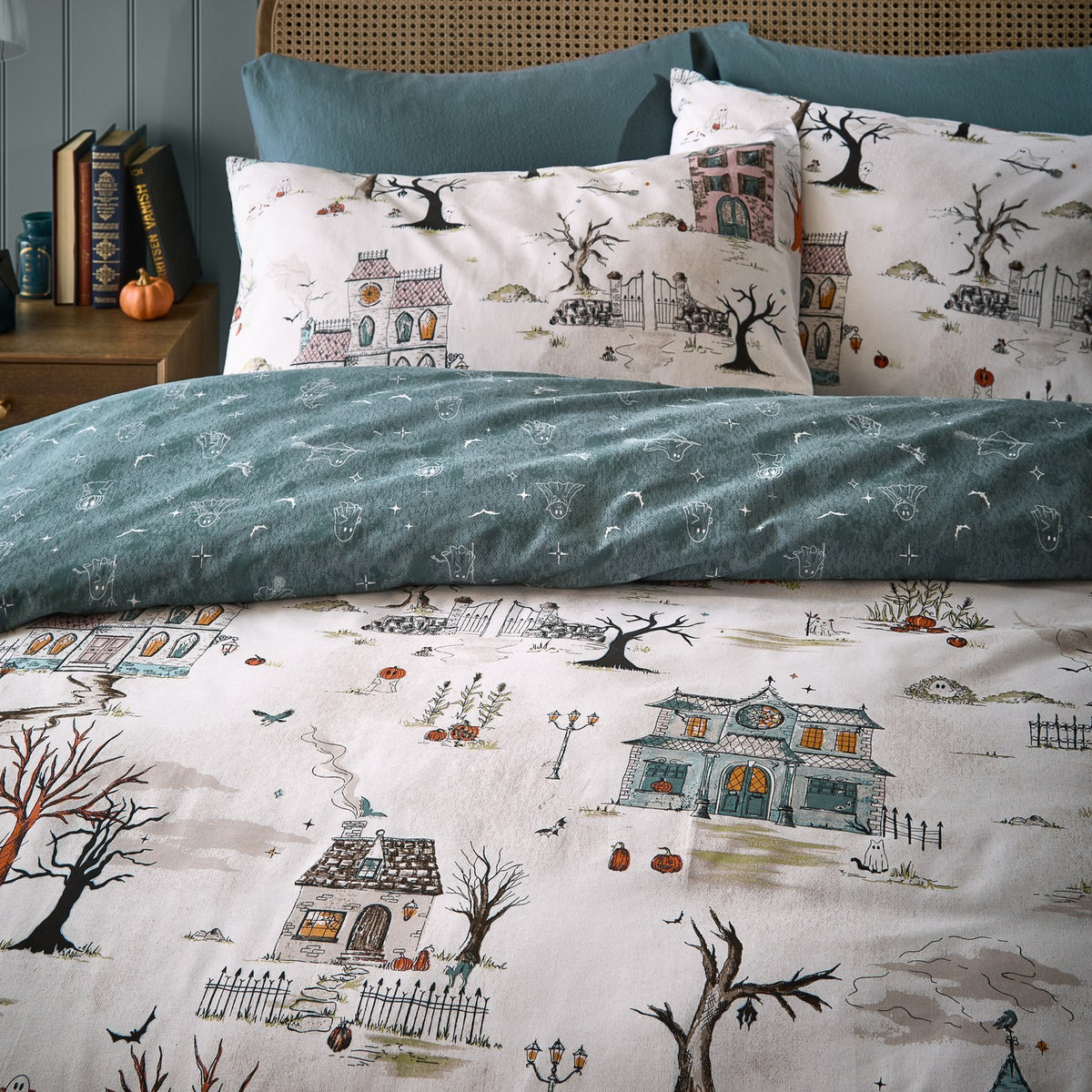 Ghost Town reversible duvet cover set on a bed, cream side showing haunted houses, trees and pumpkins, paired with dusk-blue reverse and matching pillowcases.