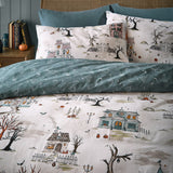 Ghost Town reversible duvet cover set on a bed, cream side showing haunted houses, trees and pumpkins, paired with dusk-blue reverse and matching pillowcases.