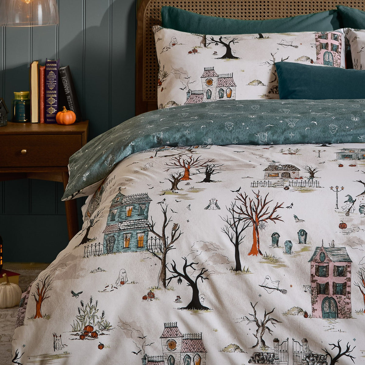 Ghost Town Reversible Woodland Duvet Cover Set in Dusk on a bed, cream side shown with haunted houses, bare trees and ghosts, teal reverse edge visible, styled bedroom setting.