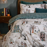Ghost Town Reversible Woodland Duvet Cover Set in Dusk on a bed, cream side shown with haunted houses, bare trees and ghosts, teal reverse edge visible, styled bedroom setting.