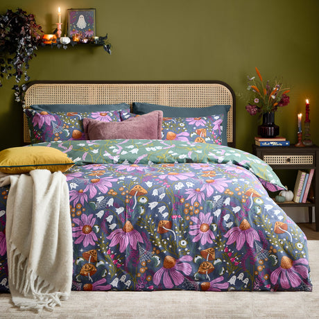 Ghoul Gardens reversible duvet cover set on a bed, slate-blue side with pink coneflowers, orange toadstools and white ghosts, styled in a green-walled bedroom with cane headboard.