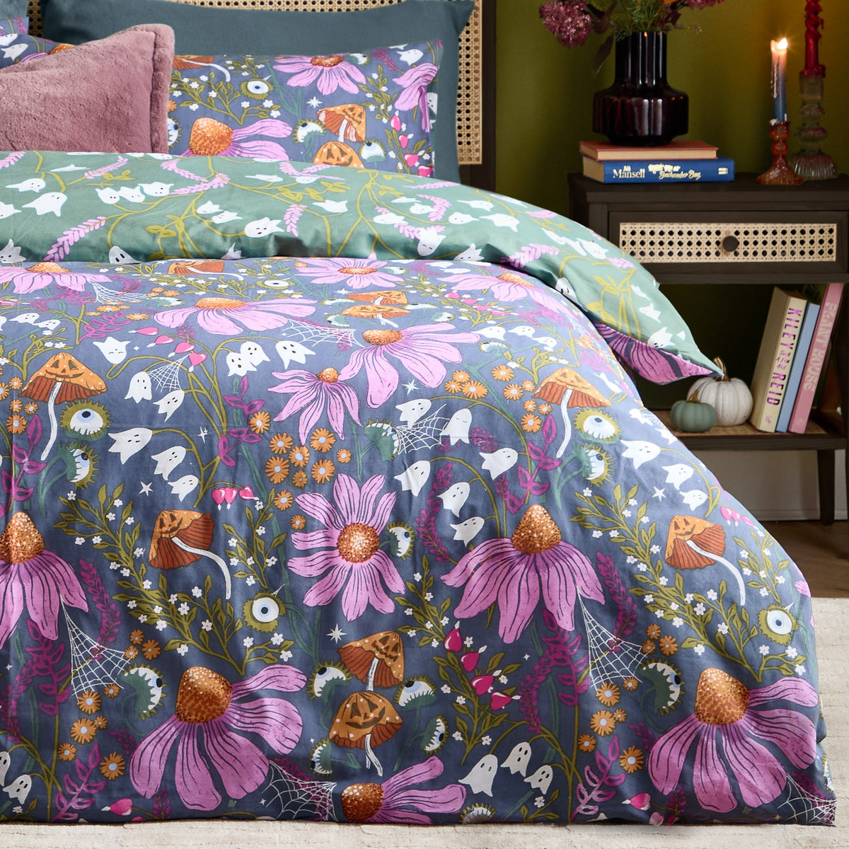 Ghoul Gardens reversible duvet cover set on a bed, slate-blue side with pink coneflowers, orange toadstools and ghosts, paired with a sage-green ghost print pillowcase.