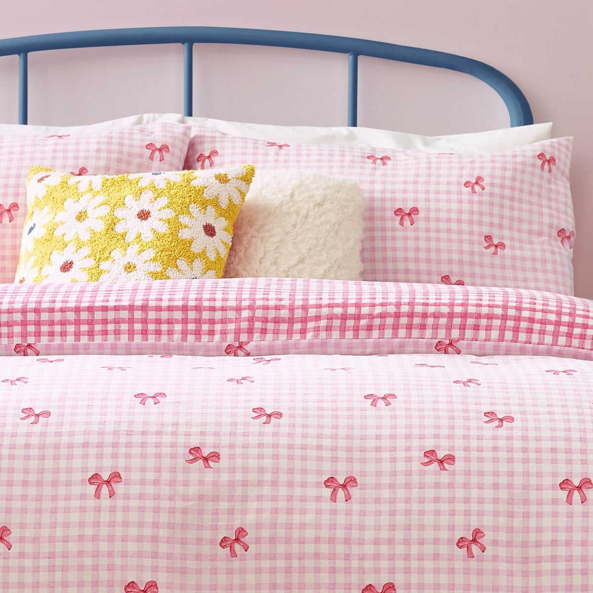 Pink gingham check duvet cover set with scattered bow print, styled on a bed with matching pillowcases, a yellow daisy cushion, and a blue metal headboard.