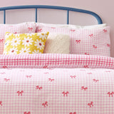 Pink gingham check duvet cover set with scattered bow print, styled on a bed with matching pillowcases, a yellow daisy cushion, and a blue metal headboard.