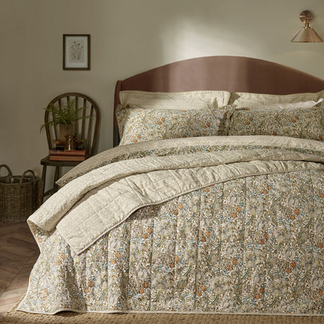 Golden Lily quilted cotton bedspread in natural and gold floral print, shown folded on a bed with matching pillowcases in a bedroom setting.