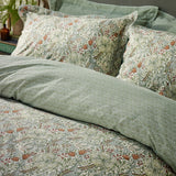 Golden Lily cotton duvet cover set in herbal green, showing swirling lily and tulip print in soft greens and rust with tonal damask reverse, styled on a bed with matching pillowcases.