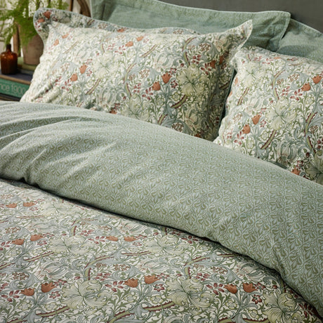 Golden Lily cotton duvet cover set in herbal green, showing swirling lily and tulip print in soft greens and rust with tonal damask reverse, styled on a bed with matching pillowcases.