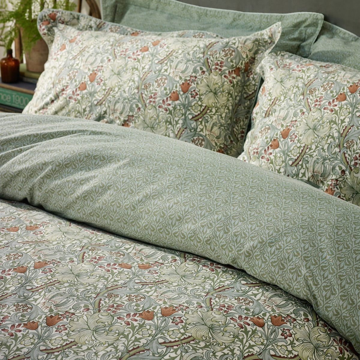 Golden Lily Percale Cotton Pillowcase Pair in Herbal Green, shown styled on a bed with matching duvet cover, featuring a classic floral pattern in green, cream, and rust tones.