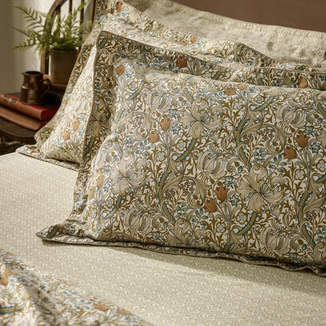 Golden Lily percale cotton pillowcases in natural and gold, shown stacked on a bedroom bed with a scrolling floral lily and tulip print and coordinating fitted sheet.