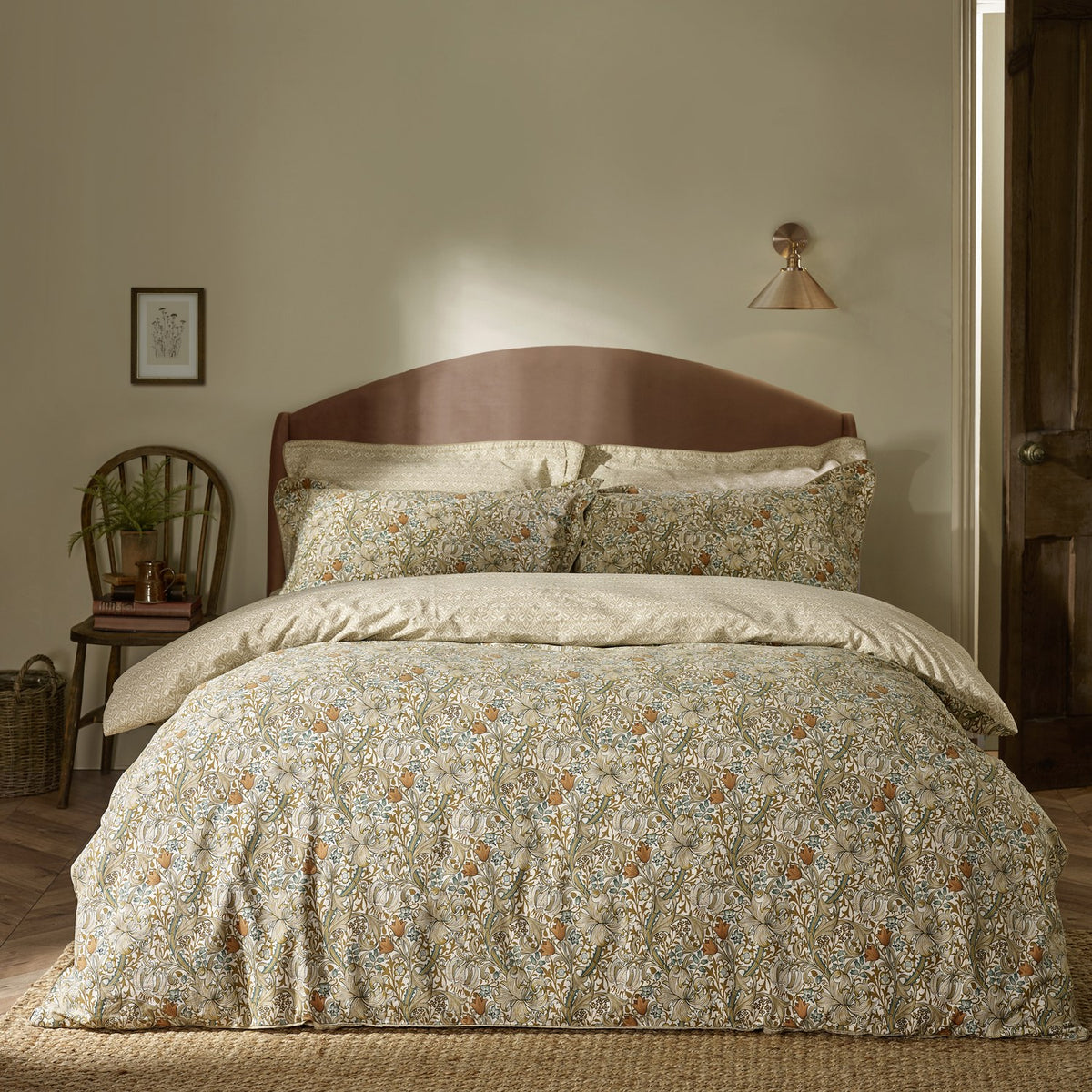 Bedroom scene with a bed dressed in Golden Lily percale cotton pillowcases and duvet set, featuring a scrolling floral lily print in natural, gold, and green tones against a wooden headboard.