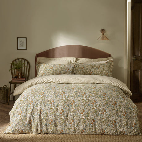 Bedroom scene with a bed dressed in Golden Lily percale cotton pillowcases and duvet set, featuring a scrolling floral lily print in natural, gold, and green tones against a wooden headboard.