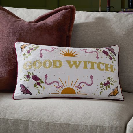 Rectangular cream velvet cushion on a sofa, featuring "Good Witch" in gold lettering surrounded by printed roses, butterflies, snakes and a sun motif, with burgundy piping.