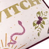 Close-up of the Good Witch velvet cushion in Angel White, showing gold embroidered lettering with a printed snake, roses, leaves and sun motif on cream velvet, edged in burgundy piping.