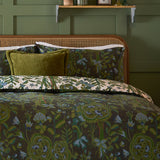Garden of Wonder Reversible Floral Duvet Cover Set in Evergreen, shown on a bed with a dark green botanical print and a cream floral reverse edge, styled with a green cushion in a rustic bedroom.