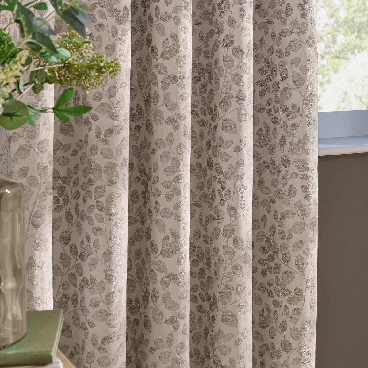 Grace Leaf Jacquard eyelet curtains in natural, showing fully lined fabric with a woven trailing leaf pattern hung in soft folds beside a window.