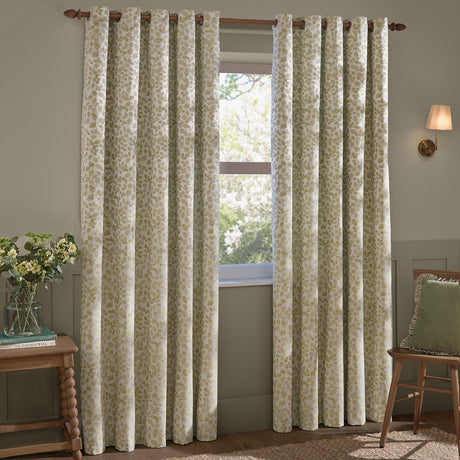 Grace Leaf Jacquard fully lined eyelet curtains in sage, cream fabric with a woven leaf pattern, hung on a wooden pole in a living room beside a window.
