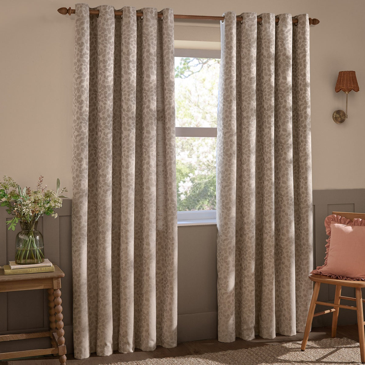 Grace Leaf Jacquard fully lined eyelet curtains in taupe with a tonal woven leaf pattern, shown hanging on a wooden pole beside a window in a living room setting.