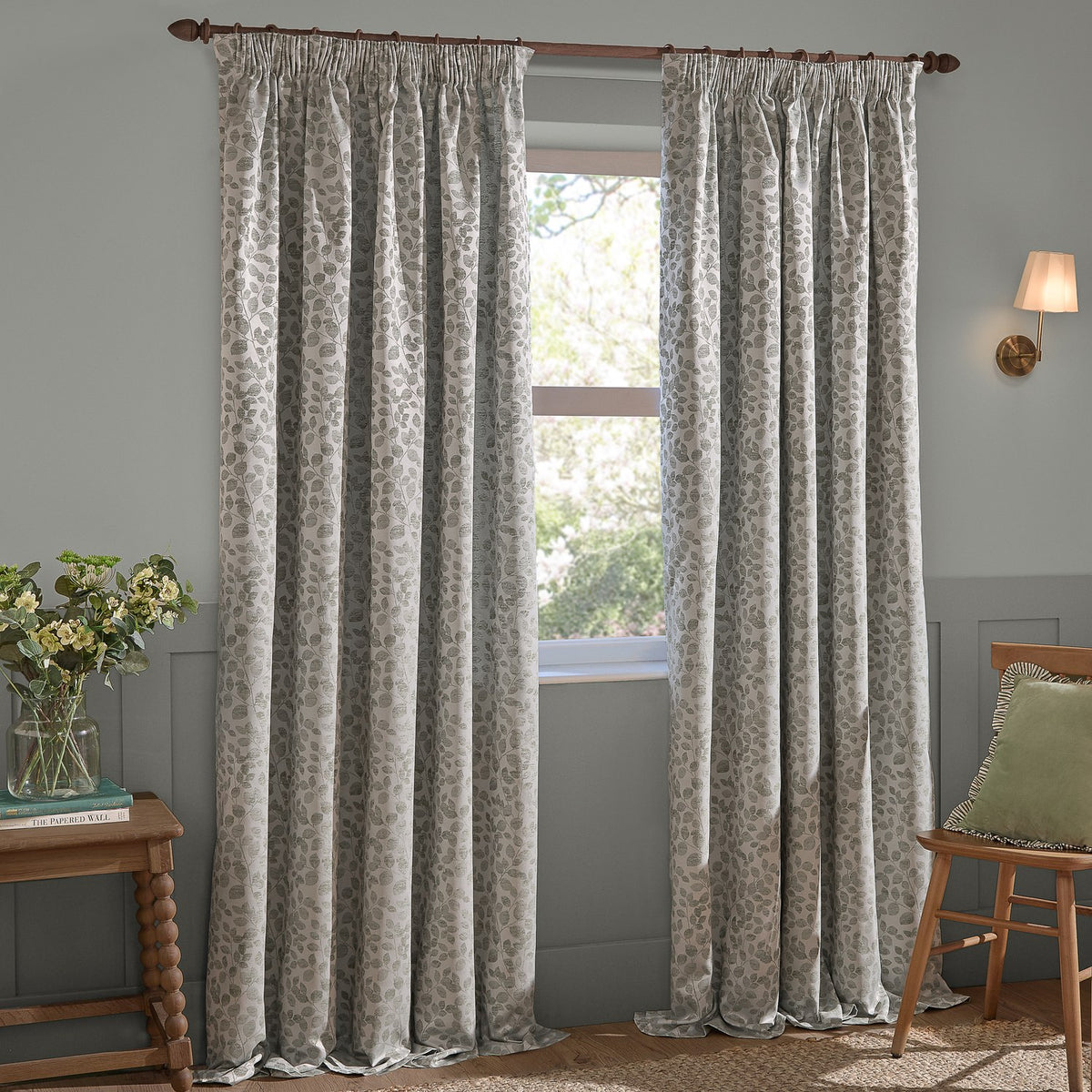 Grace Leaf Jacquard pencil pleat curtains in mist grey, shown full-length on a wooden pole in a living room with matching wall panelling and window.