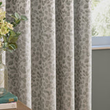 Grace Leaf Jacquard pencil pleat curtains in mist grey with woven leaf pattern, shown hanging in a pencil-pleat fold beside a window in a living room setting.