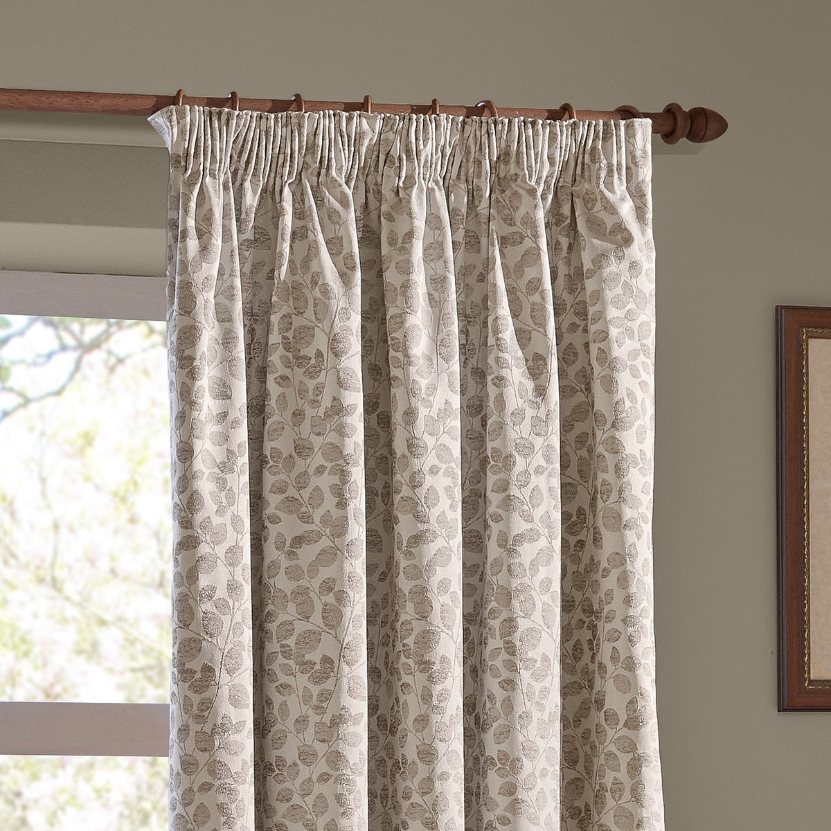 Grace Leaf Jacquard Polyester Pencil Pleat Curtains in natural beige, showing a trailing leaf jacquard pattern, hung on a wooden pole beside a window.