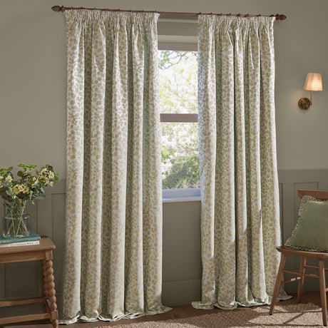 Grace Leaf Jacquard pencil pleat curtains in sage on a wooden pole, showing the leaf pattern woven in green on cream fabric in a living room setting beside a window.