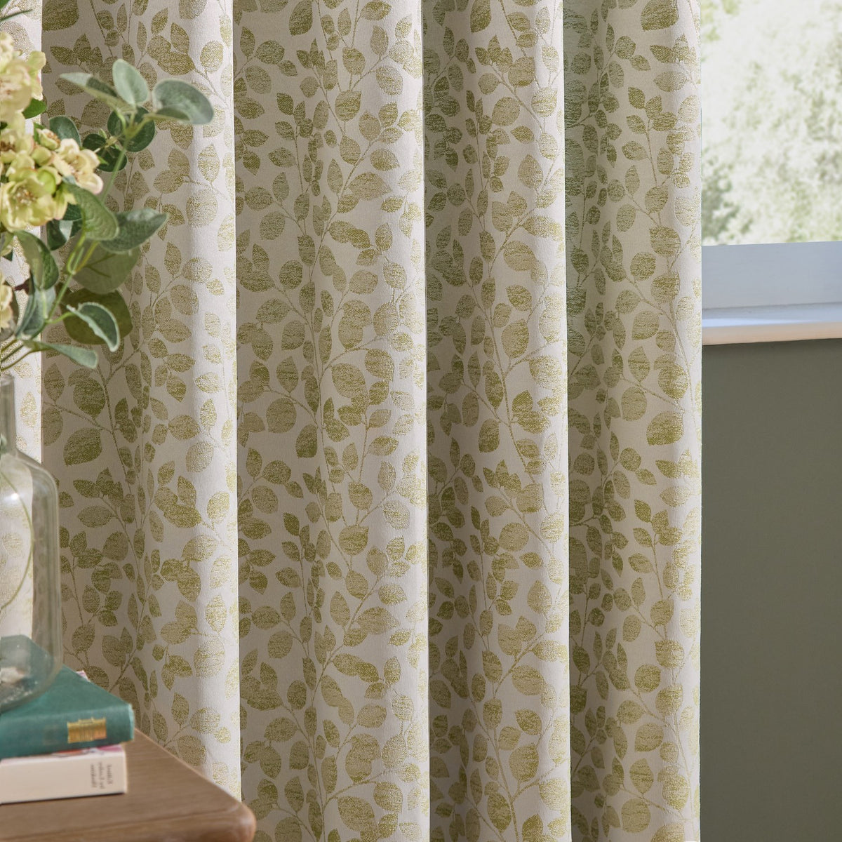 Grace Leaf Jacquard pencil pleat curtains in sage, showing cream fabric with woven leaf pattern and pleated header, styled beside a window in a living space.