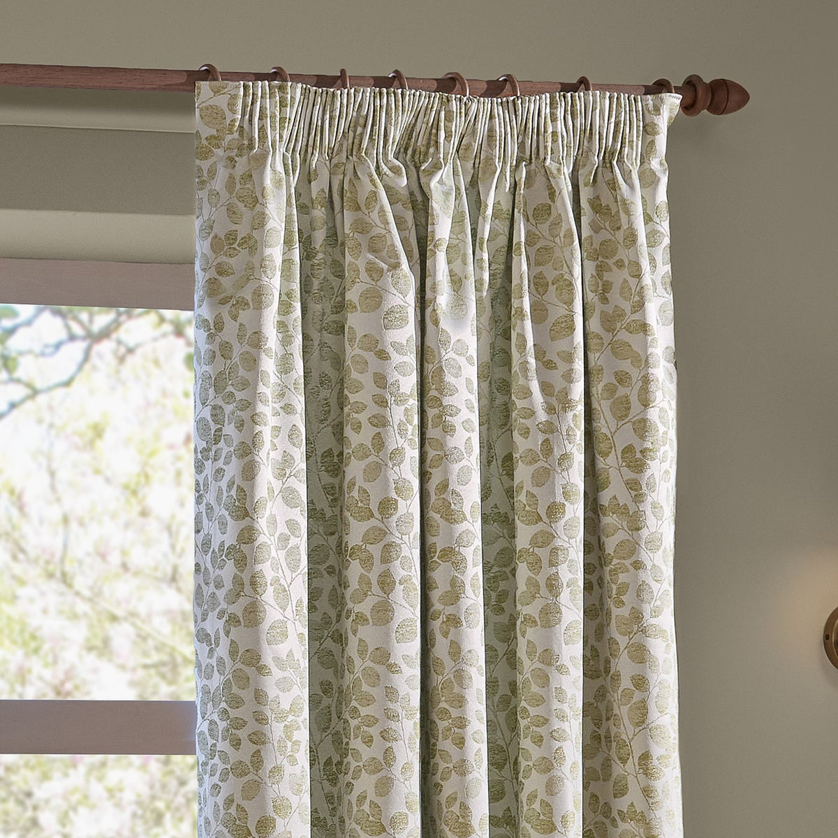 Grace Leaf Jacquard pencil pleat curtains in sage, showing a trailing leaf pattern on cream fabric, hung on a wooden pole beside a window.