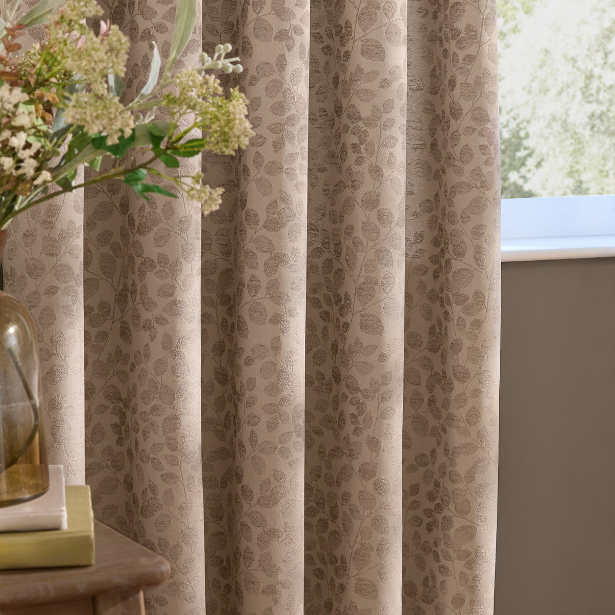 Grace Leaf Jacquard pencil pleat curtains in taupe, shown pleated by a window, with a woven leaf pattern in a matt satin polyester finish.